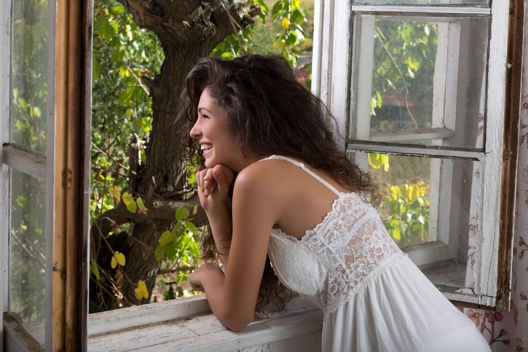 кудрявая девушка в белом платье смотрит в окно и улыбается