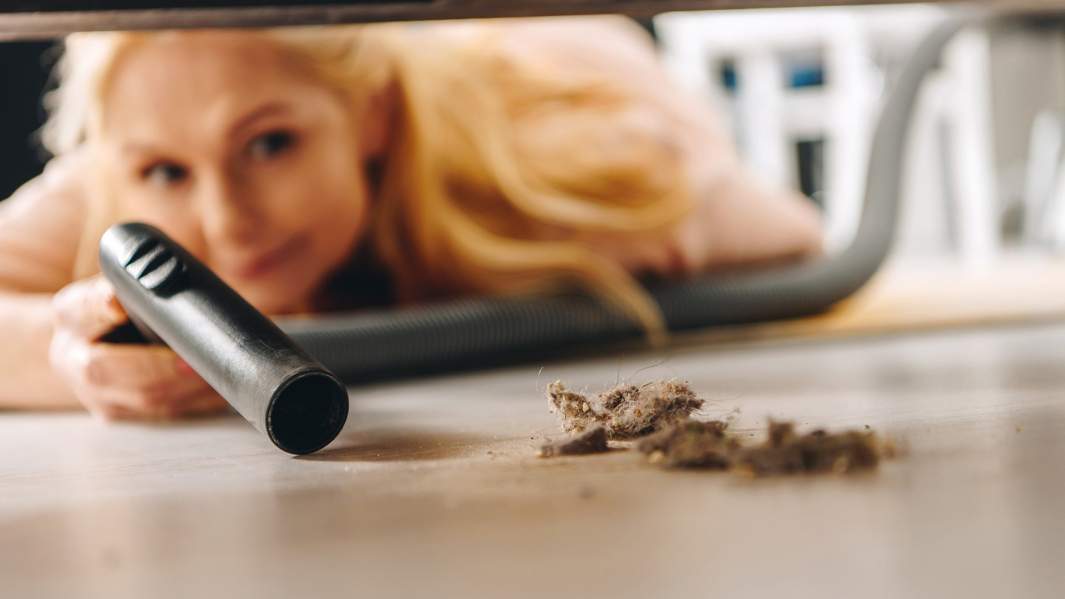 Женщина с пылесосом убирает мусор из-под дивана