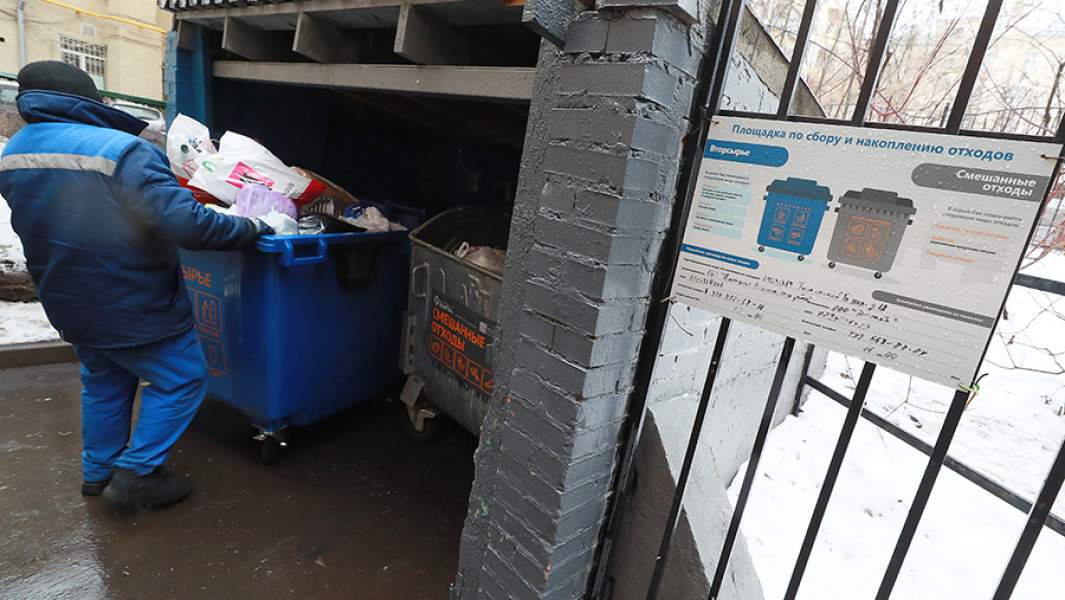 Сотрудник компании вывозит мусорный контейнер для выгрузки его содержимого в мусоровоз в одном из дворов