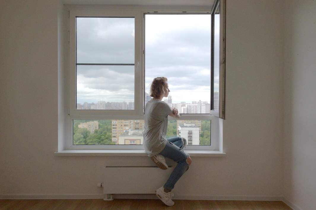 Девушка сидит на подоконнике в одной из квартир многоэтажного жилого дома