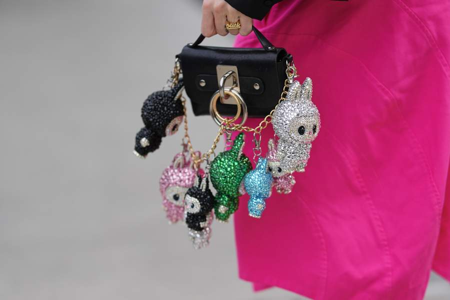 NEW YORK, NEW YORK - SEPTEMBER 12: A guest wears a hand with short natural nails wearing a gold ring with raised script, carrying a small rectangular black leather top-handle bag with a square silver metal plate and oversized round gold and silver O-ring hardware, from which a gold chain suspends multiple Labubu crystal- and rhinestone-encrusted character-shaped charms in silver, green, blue, pink and black, paired with a bright fuchsia-pink smooth woven garment with visible seam